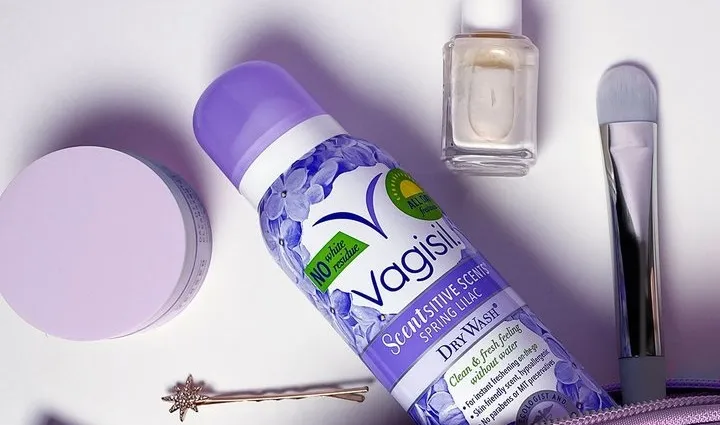 A collection of personal care items, including a purple bottle of Vagisil dry wash, a nail polish bottle, a makeup brush, a small round container, a hairpin, and a part of a makeup bag.